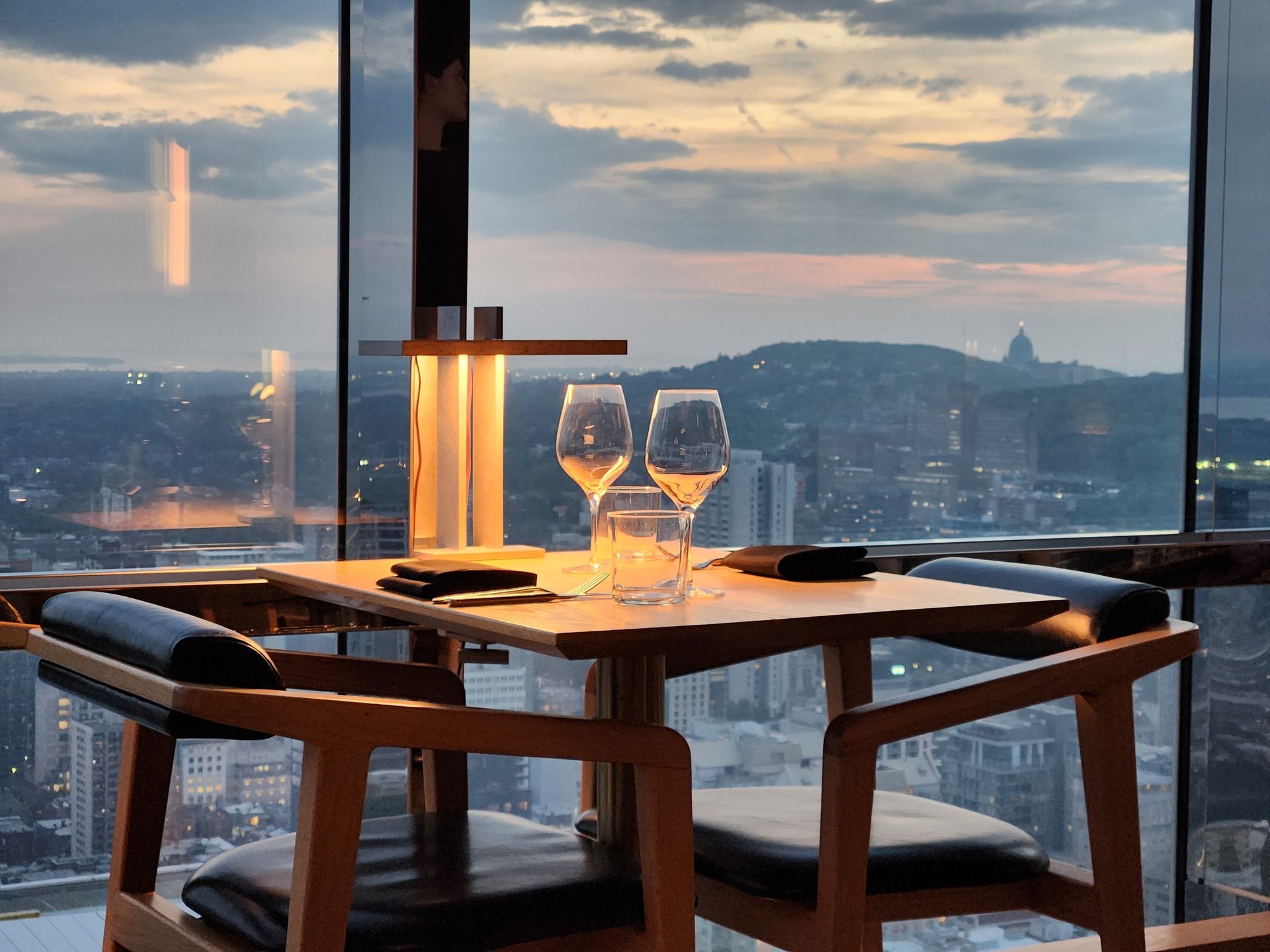 Elegant restaurant table set with wine glasses and soft ambient lighting near a large window overlooking a city skyline at sunset. The warm interior contrasts with the cool tones of the evening sky, creating a refined atmosphere ideal for concepts of fine dining, romance, luxury lifestyle, hospitality, and urban living.