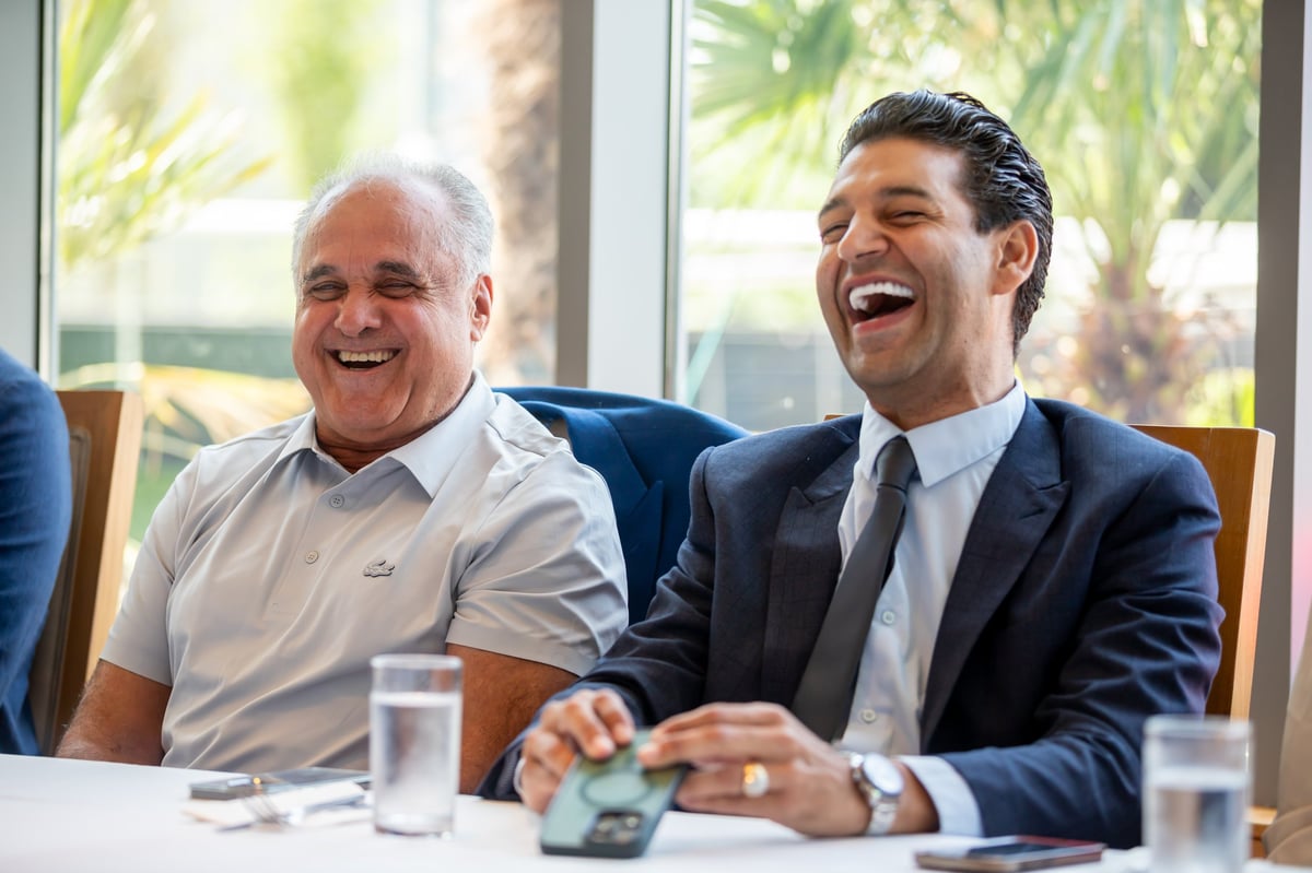 Two men laughing during a business meeting at a table with water glasses and tropical views visible through windows behind them