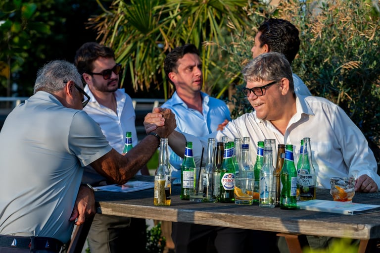 Four men in casual attire socializing outdoors around a wooden table with beer bottles in natural daylight