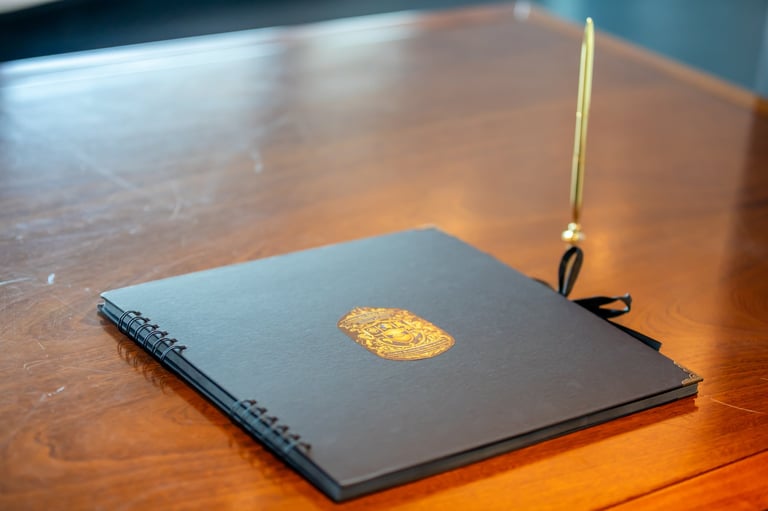 Black spiral-bound notebook with gold emblem on wooden desk next to brass pen holder with pen