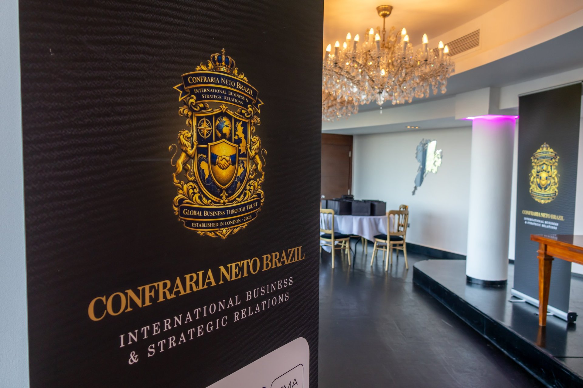 Modern office entrance with Confraria Nero Brazil signage on dark wall, crystal chandelier overhead, and elegant dining area visible in background