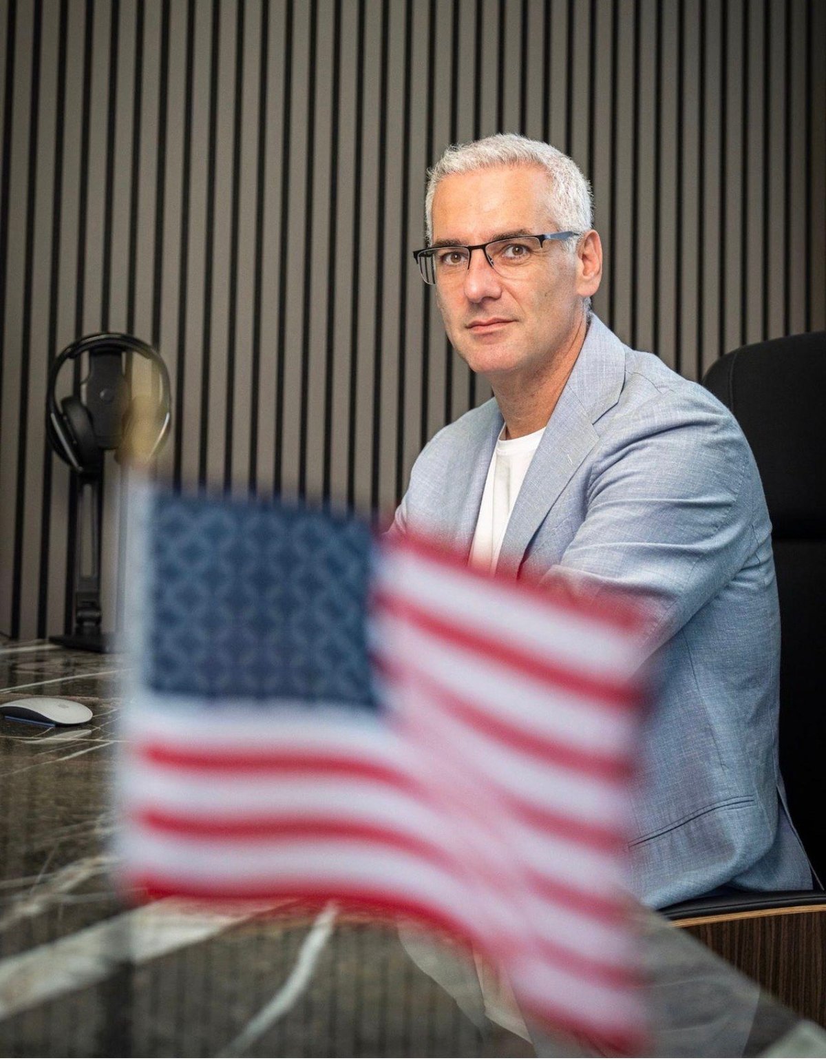 Man with white hair and glasses in light blue blazer sits at desk with American flag in foreground and microphone in background