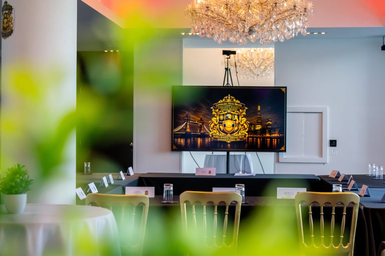 Modern dining room with mounted TV displaying illuminated architecture, crystal chandelier, wooden chairs, and plants in foreground.