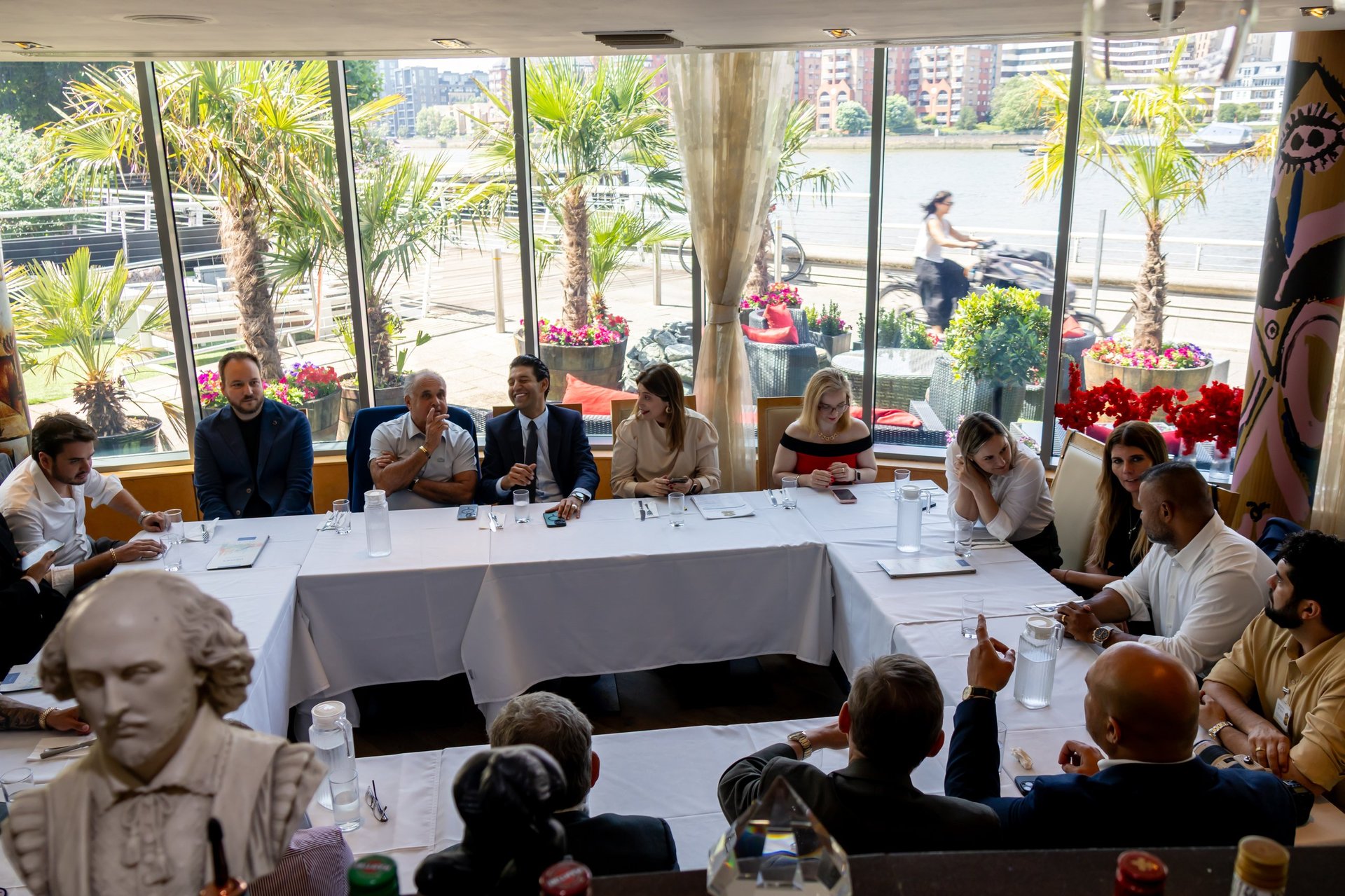Group of people seated around a conference table with large windows overlooking a palm-lined street in the background