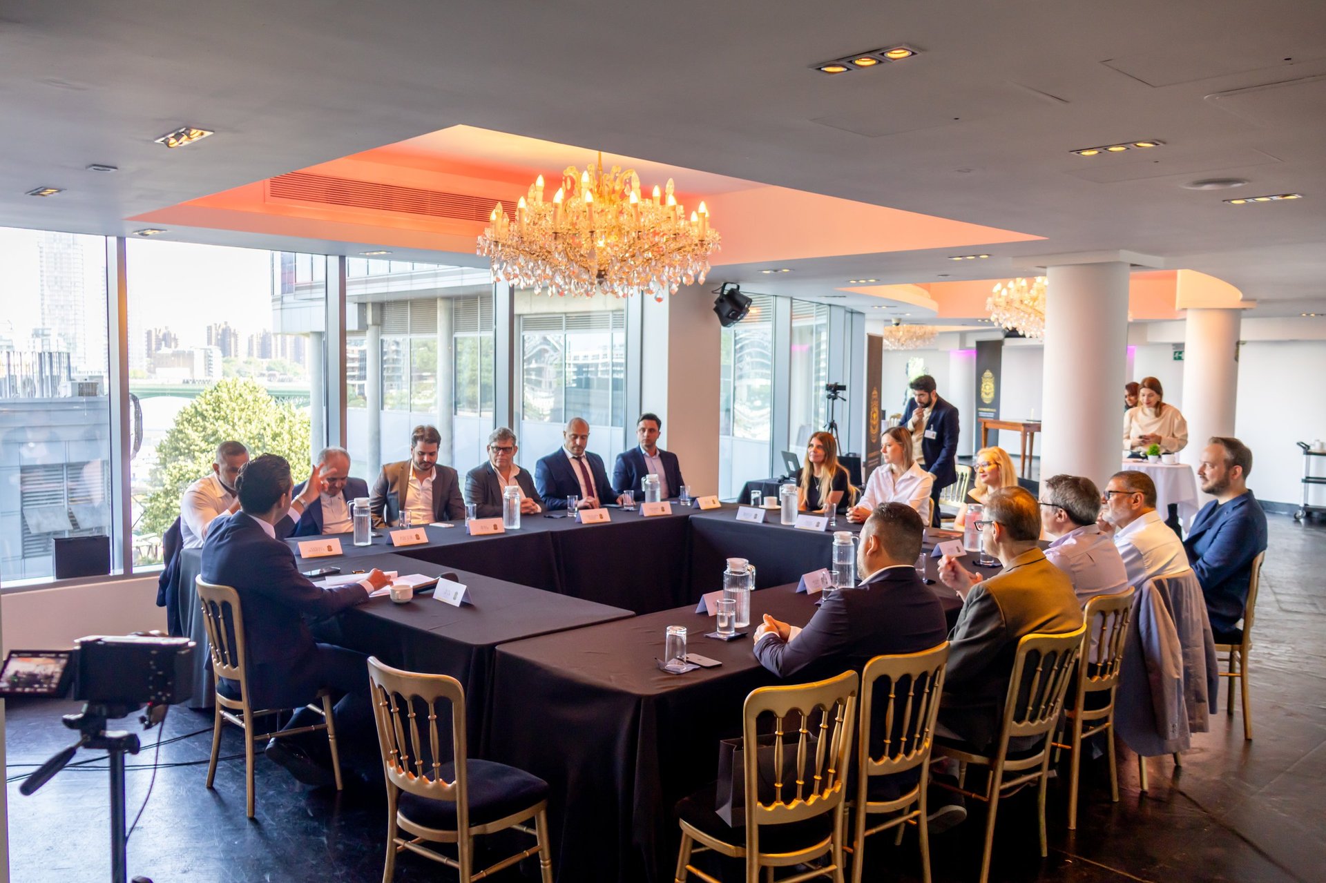 Business conference in modern office with executives seated around a black U-shaped table and standing attendees in suits