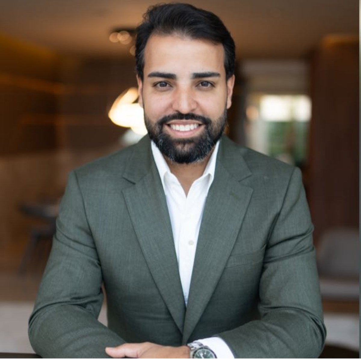 Professional man with dark beard wearing gray blazer and white shirt smiling at camera in office setting