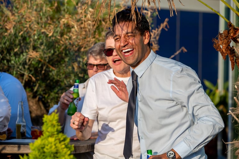 Man in light blue shirt and dark tie laughing at outdoor garden event with other guests and hanging plants in background