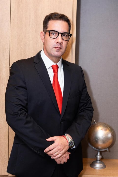 Professional man in black suit and red tie wearing glasses, standing with clasped hands next to a desk globe