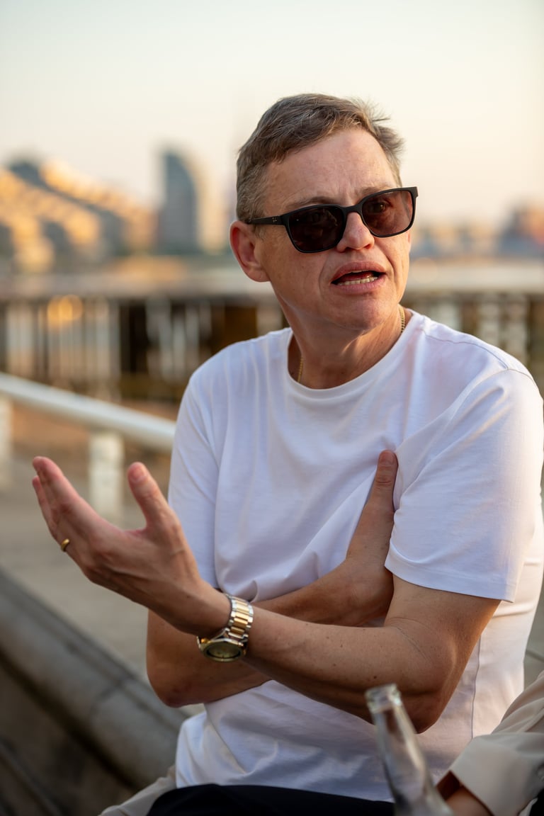 Man wearing sunglasses and white t-shirt gesturing while speaking with city skyline and bridge visible in background
