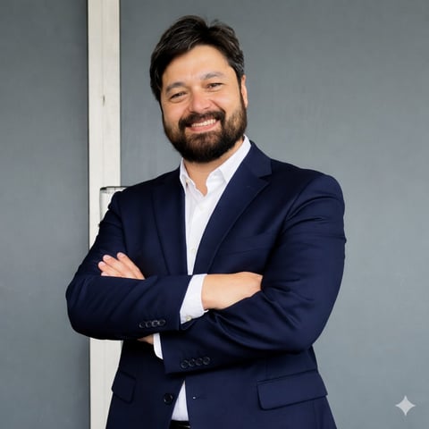 Man with beard wearing navy blazer and white shirt, arms crossed, smiling at camera against gray wall