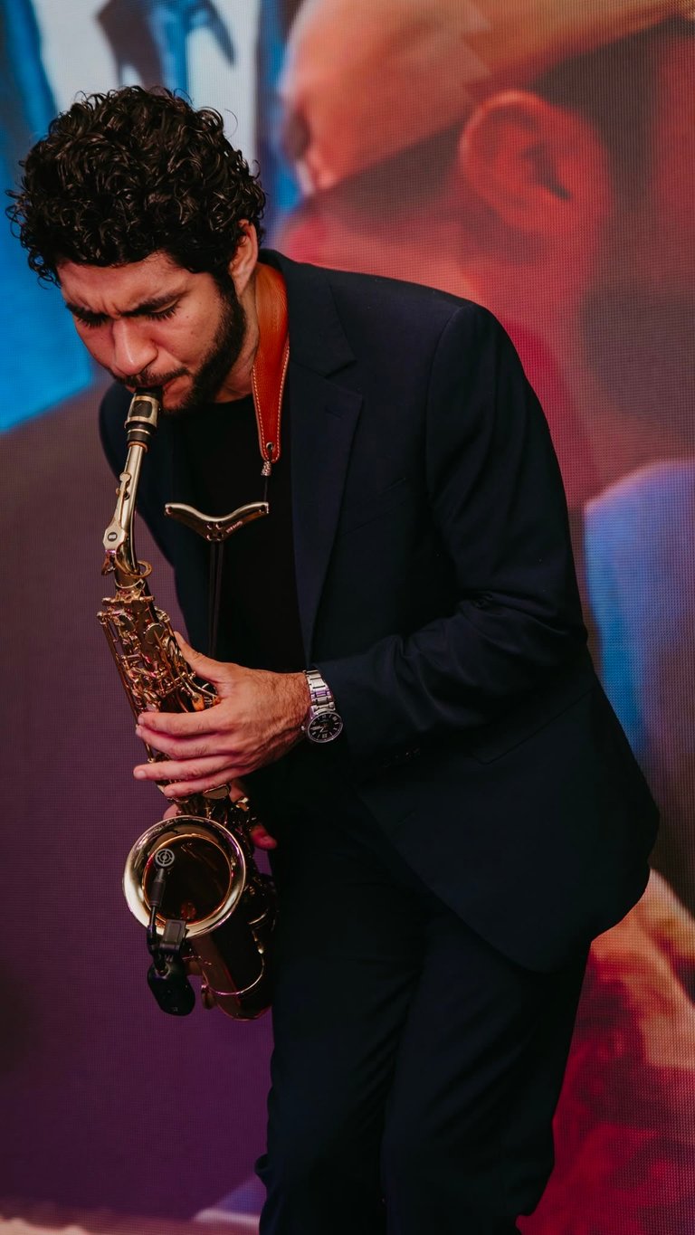 Man in black suit playing saxophone on stage with blue and red gradient lighting background
