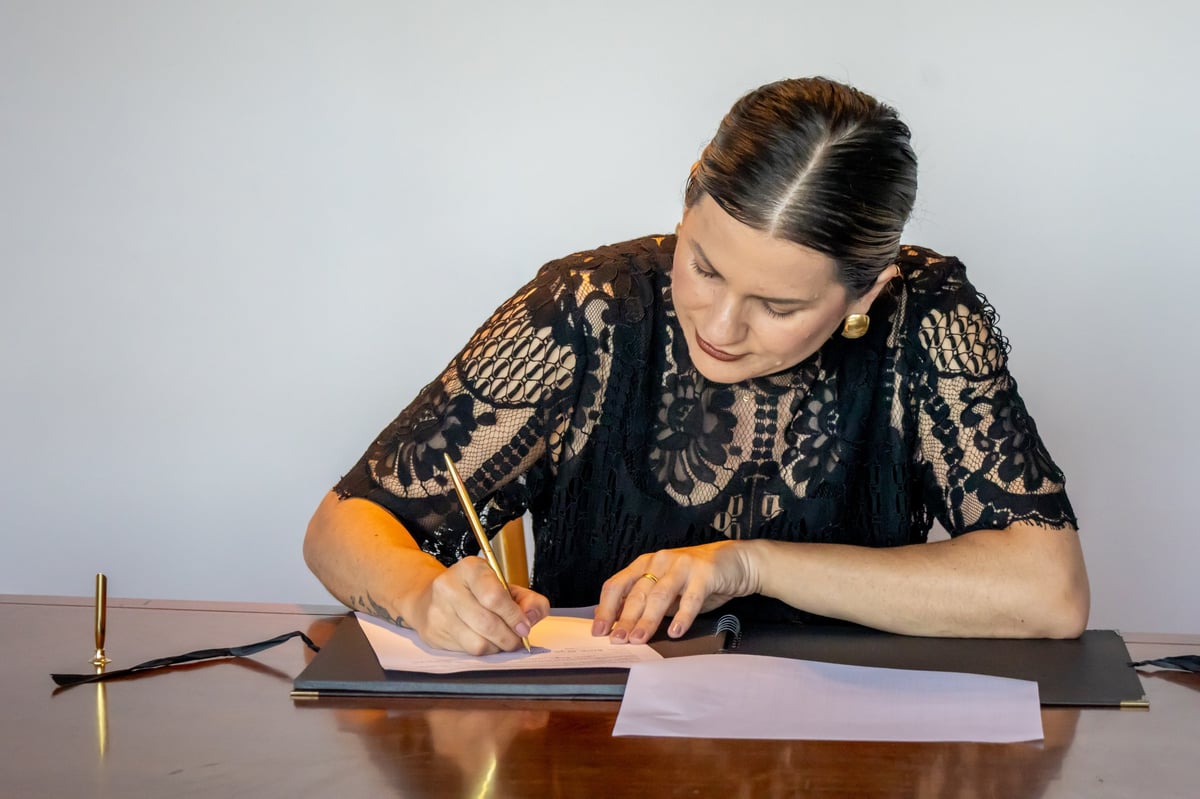 Woman in black patterned top writing on paper at desk with pen and lamp