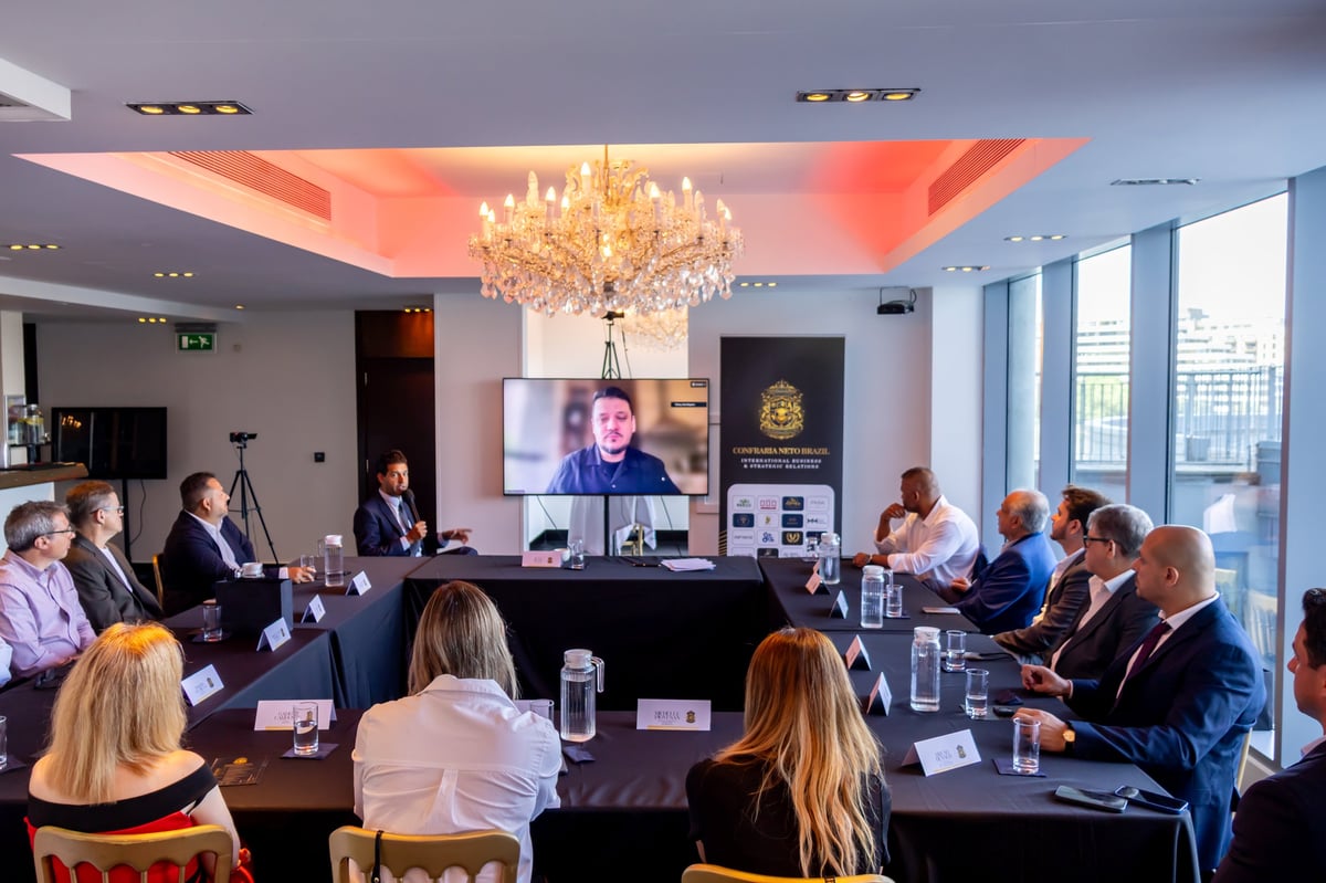 Business conference room with professionals seated around a table, watching a speaker on a large screen, with a modern chandelier overhead