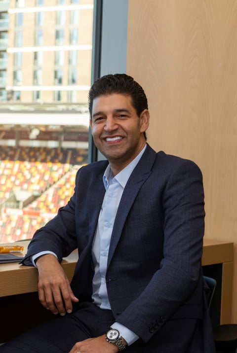 Professional man in dark blazer and light blue shirt smiling at camera near window with city view