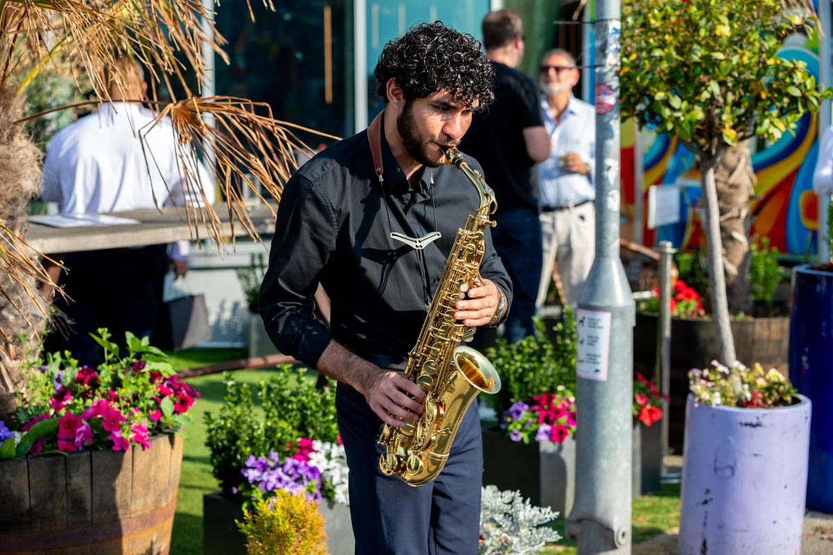 Man in black shirt playing saxophone outdoors surrounded by colorful flowers and potted plants