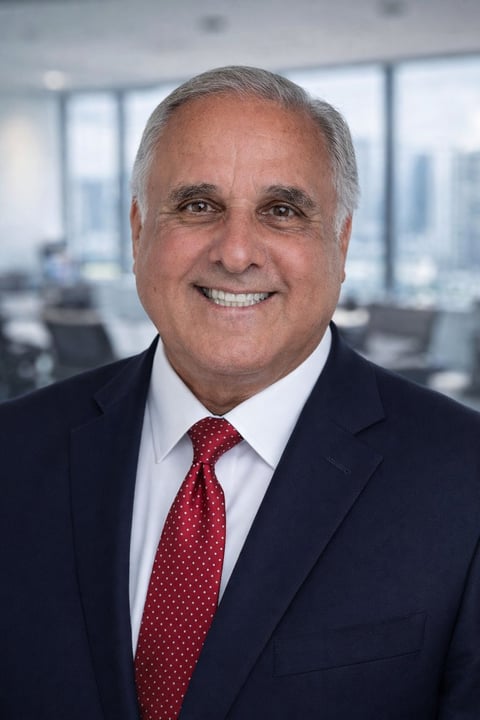Professional headshot of a middle-aged man in dark suit and red polka dot tie smiling at camera with office windows in background