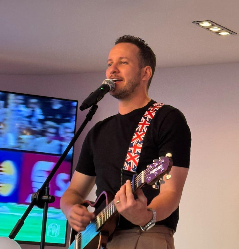 Man performing on stage with acoustic guitar and microphone, wearing black shirt with red patterned strap, colorful screens visible in background