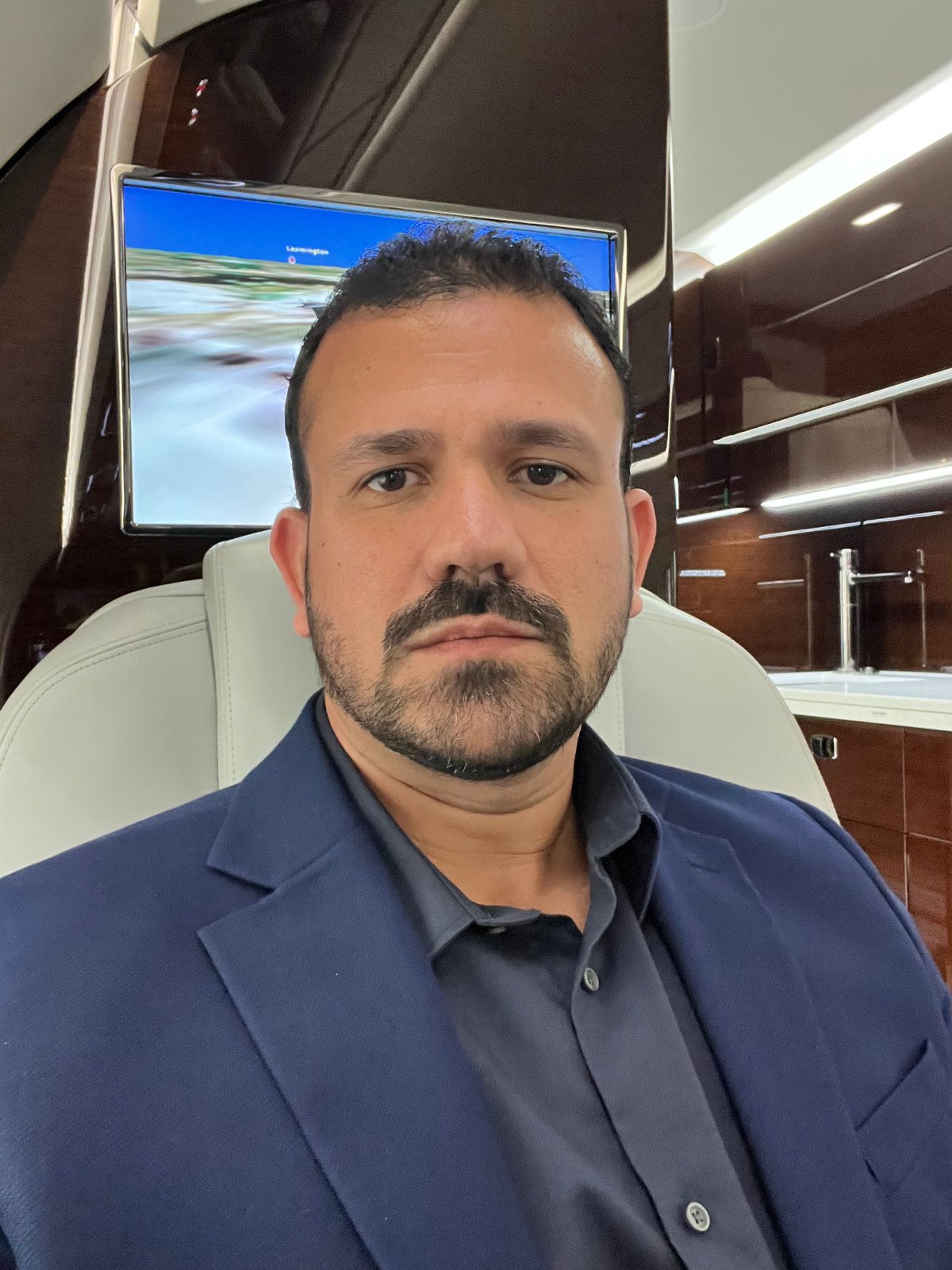Man with dark hair and beard wearing a navy blue shirt, sitting in what appears to be a luxury vehicle interior with a monitor visible in the background