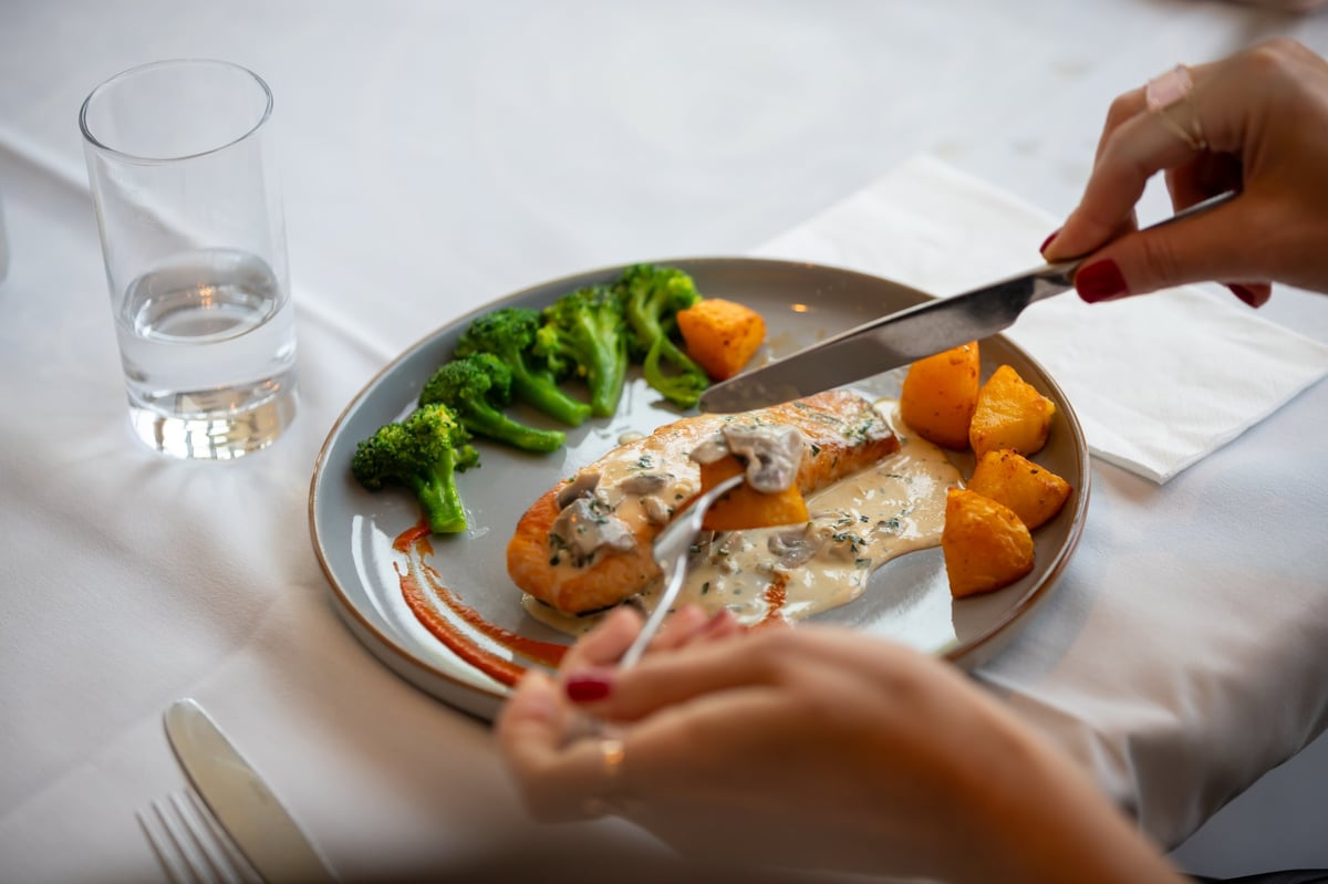 Person dining with fork over plate of grilled fish, roasted potatoes, and broccoli with water glass on white table