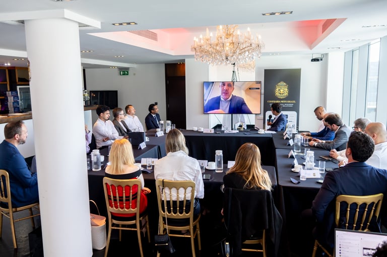 Group of people in formal business attire seated at a conference table with a large screen displaying a speaker during a virtual meeting