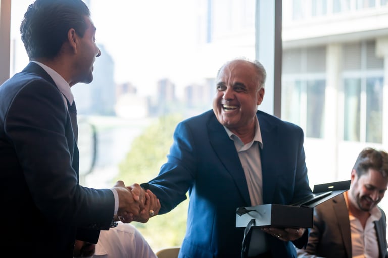Two businessmen in suits shaking hands during a meeting in a high-rise office with city skyline visible in background