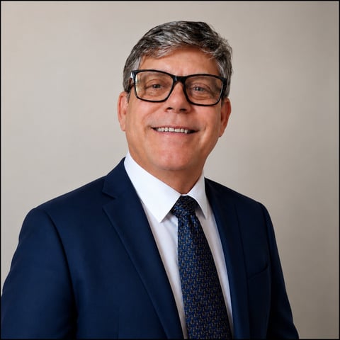 Smiling man with gray hair wearing glasses, navy blazer, and blue tie against a neutral background
