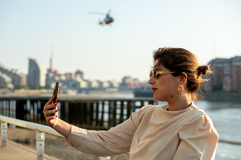 Woman in sunglasses taking a selfie with a drone flying above her by a waterfront with city buildings in the background