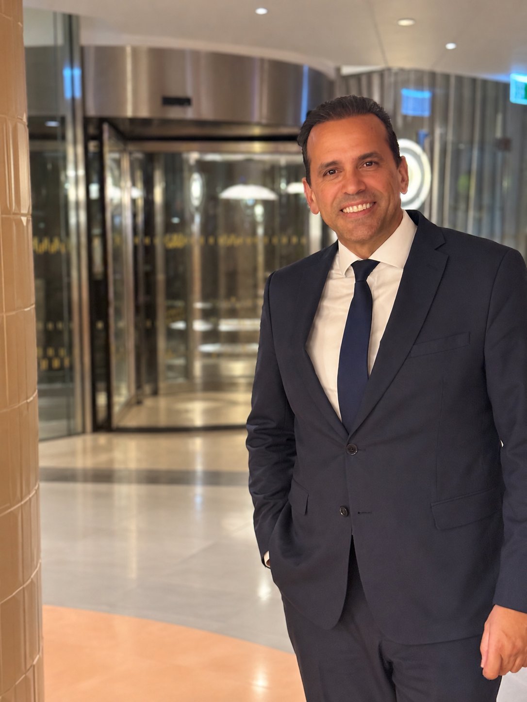 Professional man in navy suit and tie smiling in a modern office lobby with glass doors and contemporary lighting