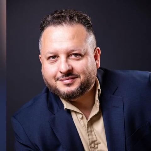 Professional headshot of a bearded man in a dark blazer and light shirt against a dark background