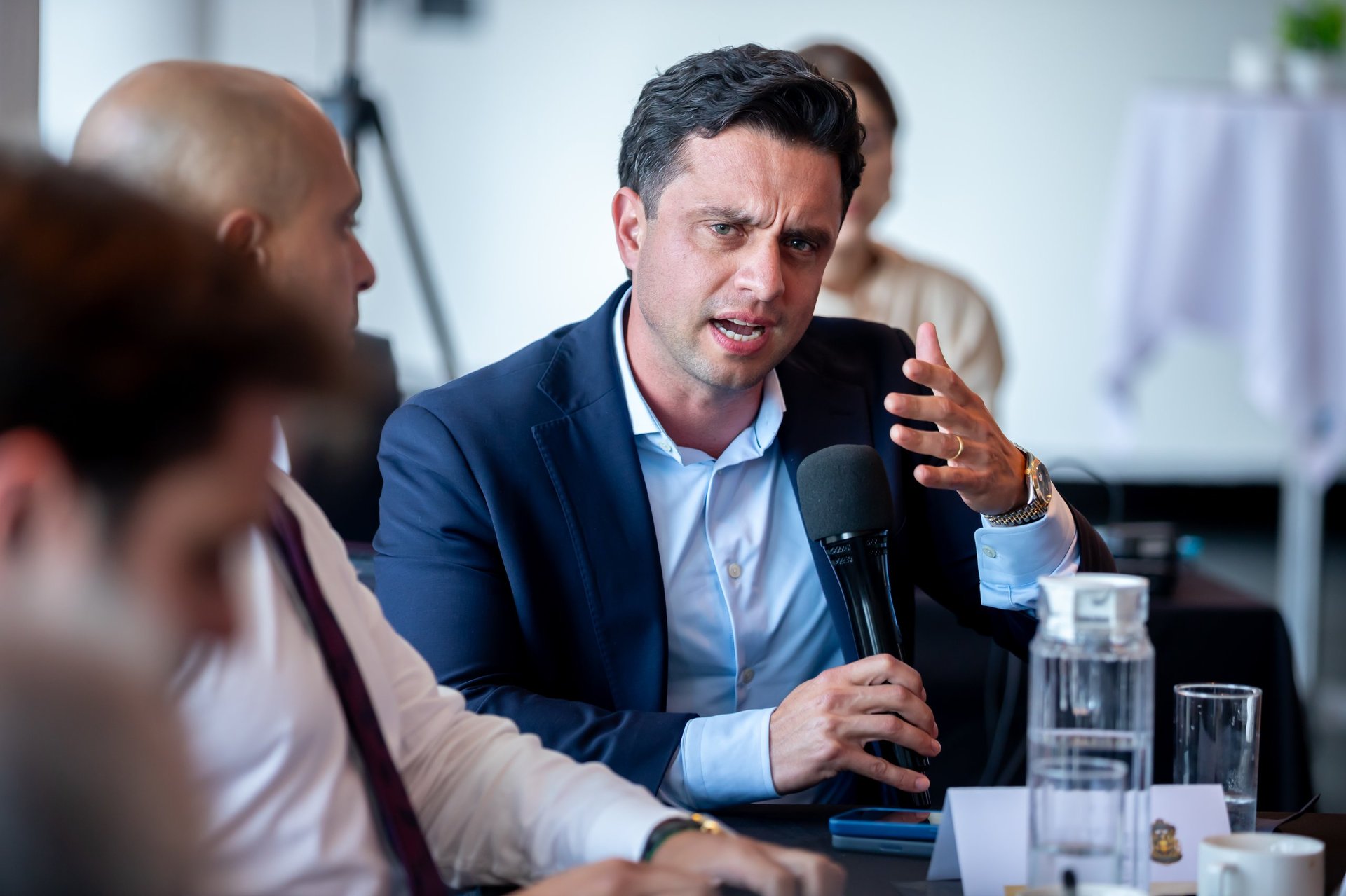 Man in navy blazer speaking at a business meeting or conference, holding a microphone while gesturing to other attendees.