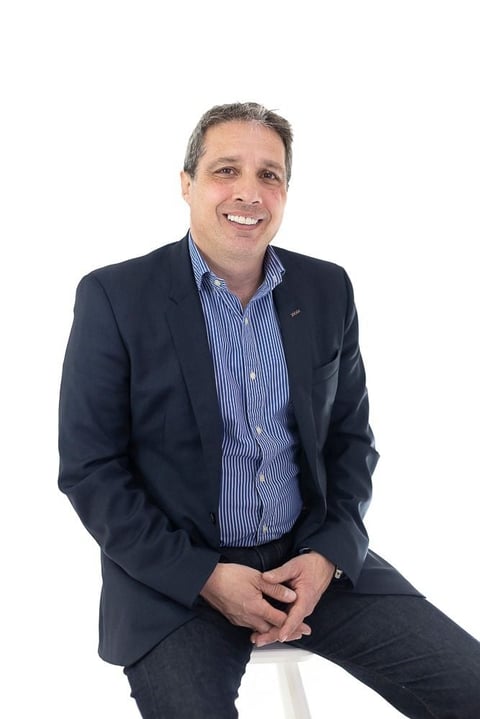 Professional headshot of a smiling man wearing a dark blazer and blue striped shirt, sitting against a white background