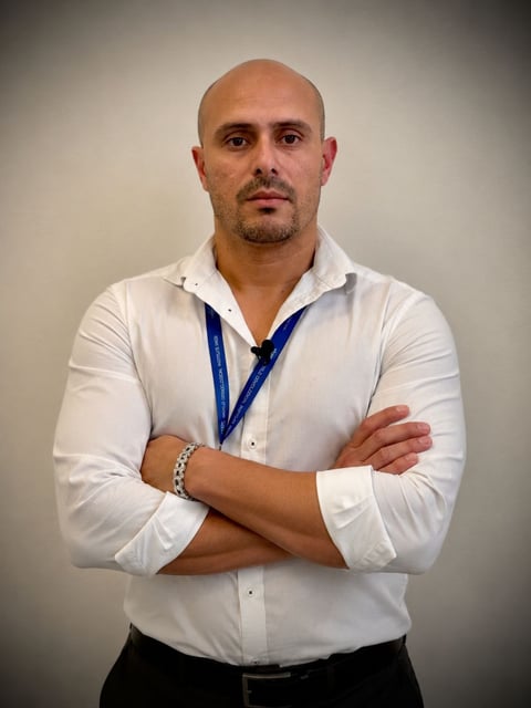Bald man in white shirt with blue lanyard, arms crossed, looking at camera against neutral background