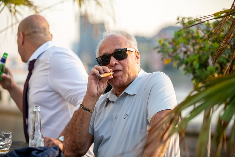 Older man wearing sunglasses and light blue shirt smoking cigar at outdoor gathering with other people and greenery visible