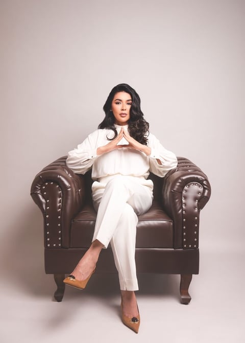 Woman in white suit sitting on brown leather armchair with hands positioned at chest, against neutral background
