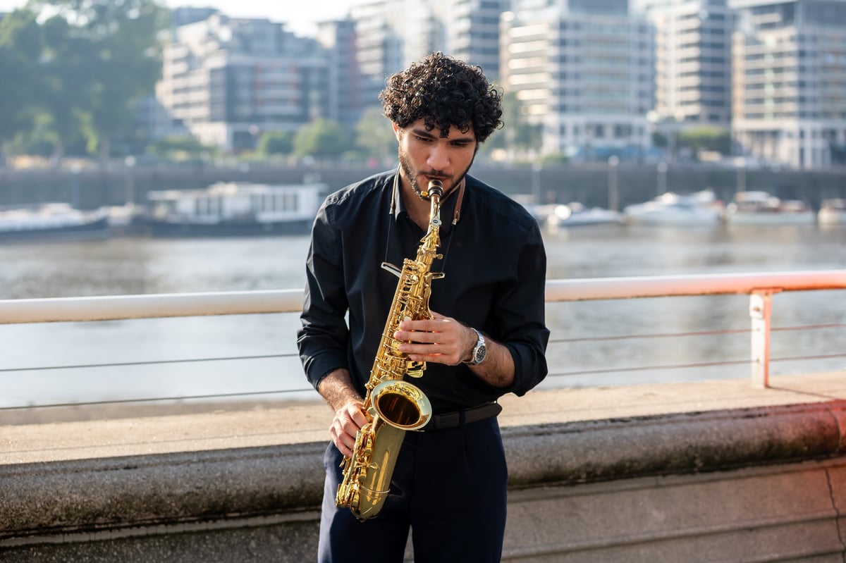 Man playing golden saxophone on urban waterfront with riverside buildings in the background