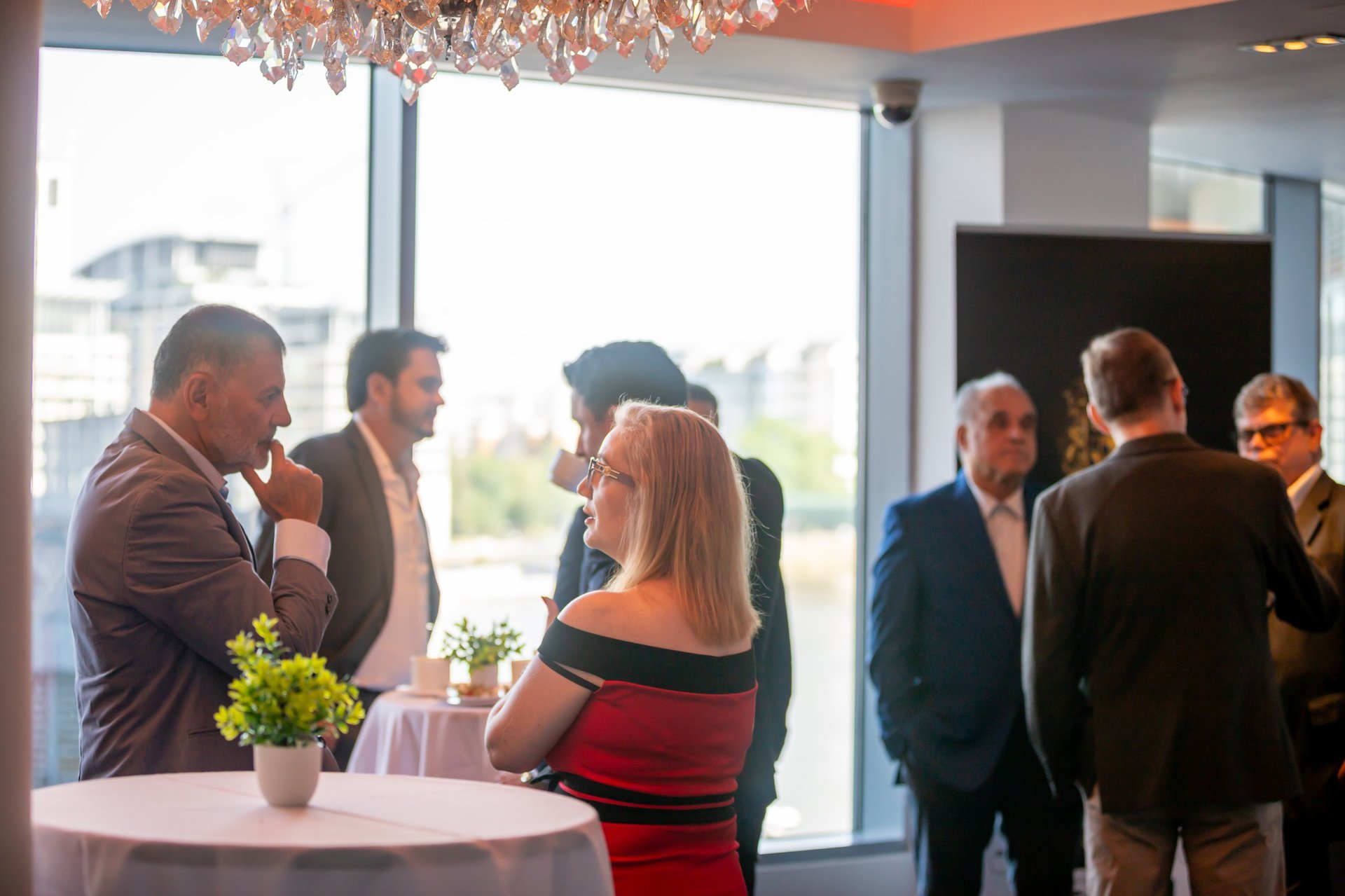 Group of professionals mingling at an indoor event near large windows, with one woman in a red and black off-shoulder dress in the foreground
