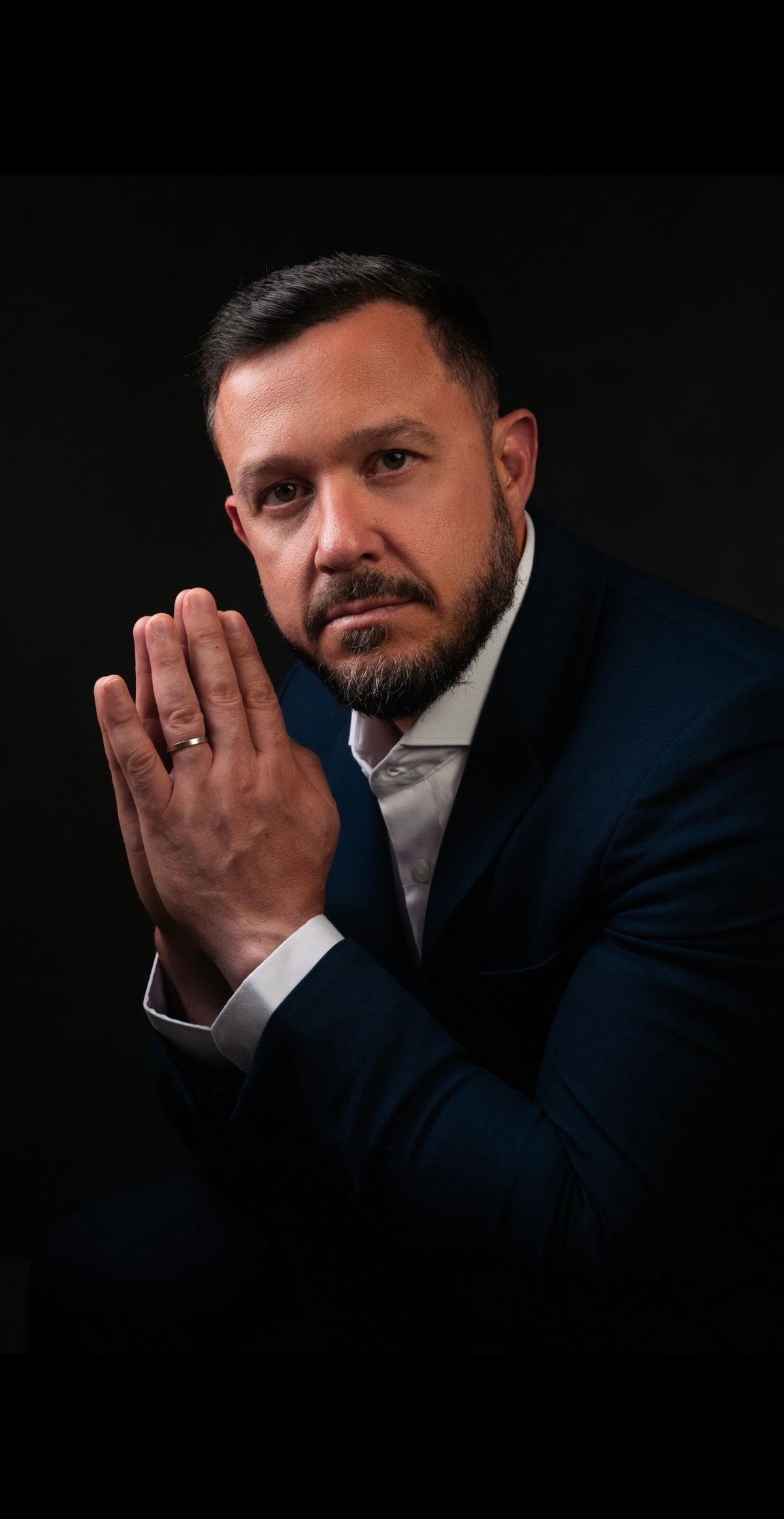 Professional man in dark suit with beard raises hand in a stopping gesture against black background