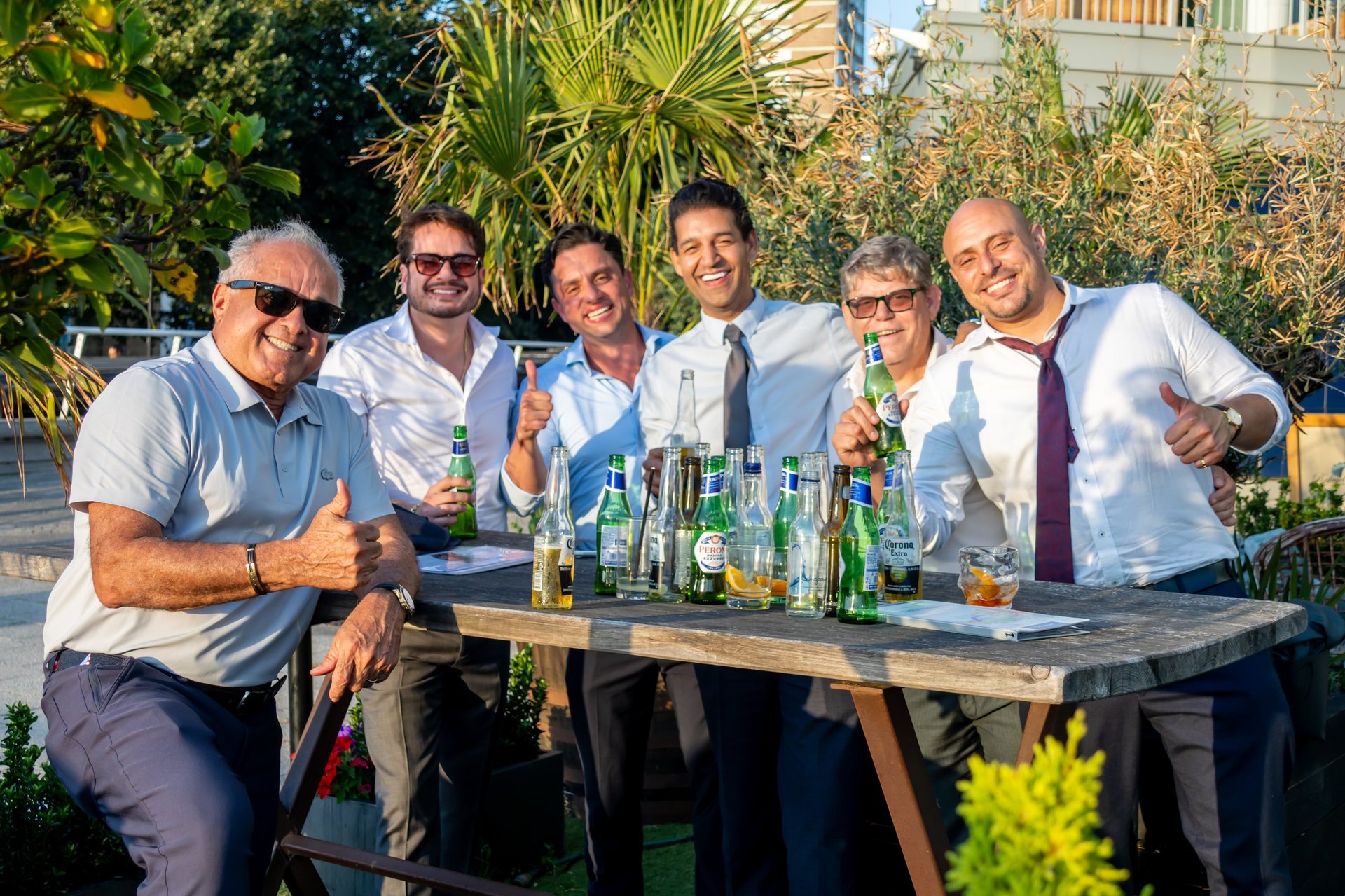 Six men in casual and business casual attire posing behind a table with beer bottles, smiling outdoors in sunlight with tropical plants and buildings visible in background