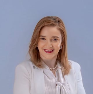Professional headshot of a woman with shoulder-length reddish-brown hair wearing a white blouse against a light blue background