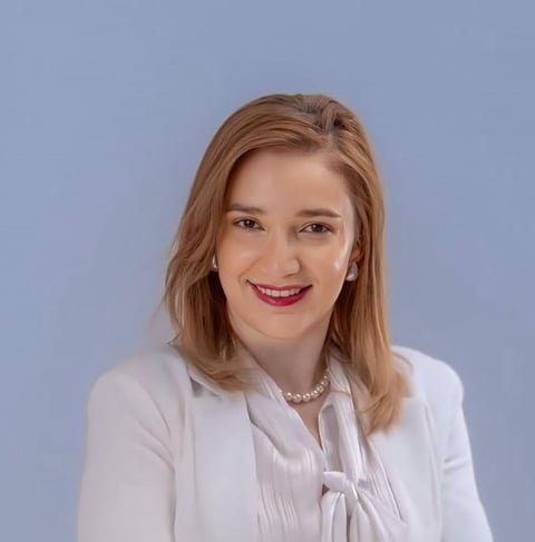 Professional headshot of a woman with shoulder-length reddish-brown hair wearing a white blouse against a light blue background