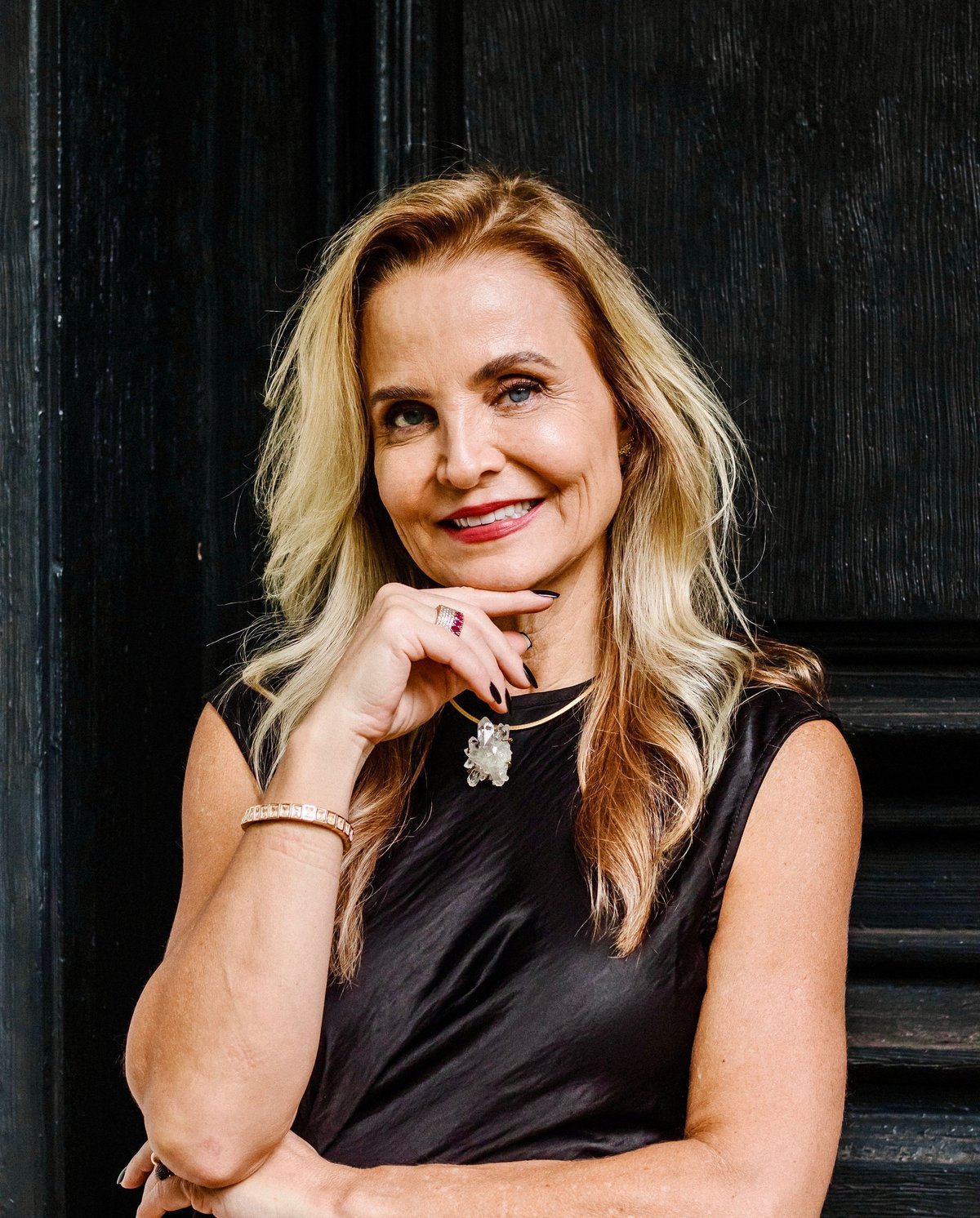 Woman with long blonde hair wearing a black sleeveless top and jewelry, smiling at camera with hand near chin against dark textured background