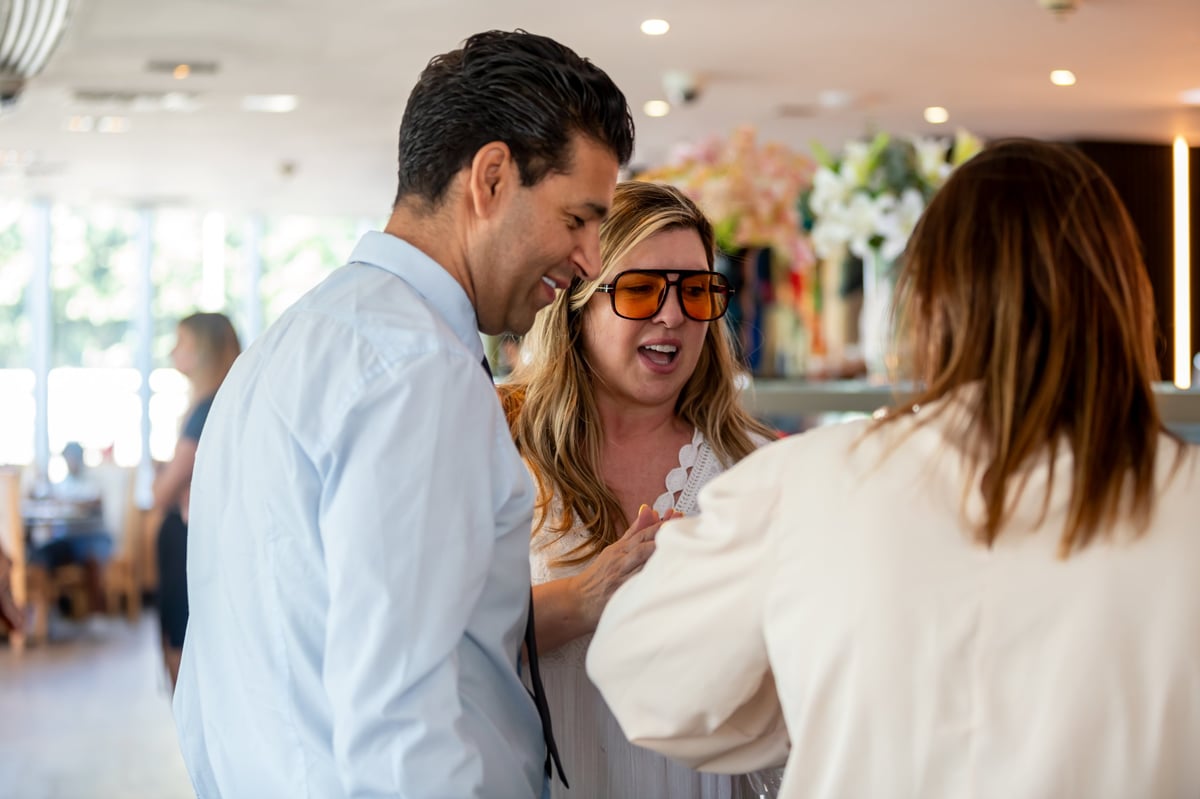 A man in a light blue shirt and a woman wearing sunglasses and a beige sweater chat with another woman in a modern, well-lit indoor venue