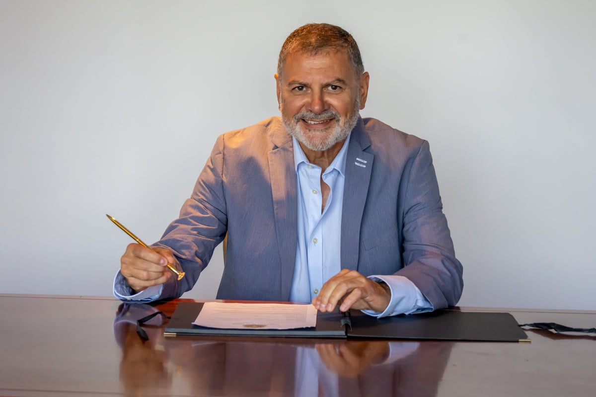 Middle-aged man in blue blazer and light blue shirt smiling at desk while holding pen over document
