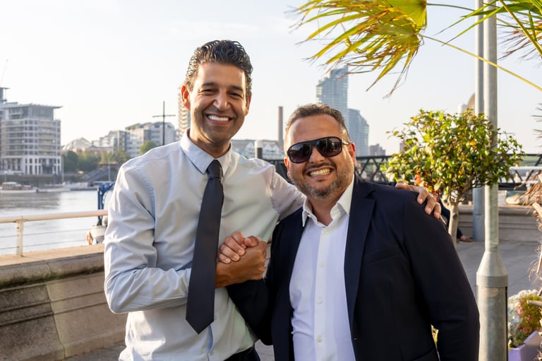 Two men in business attire smiling at camera on waterfront with city skyline in background